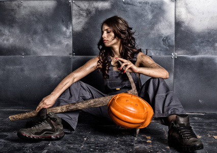 美丽疲惫的矿工工人在橙色头盔与手电筒坐在地板上的钢铁背景。手持镐的妇女