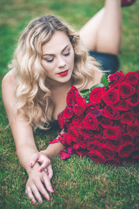 优雅美丽的金发女郎的肖像穿着黑色的时尚礼服躺在绿色的草坪上, 捧着101朵红玫瑰的大花束