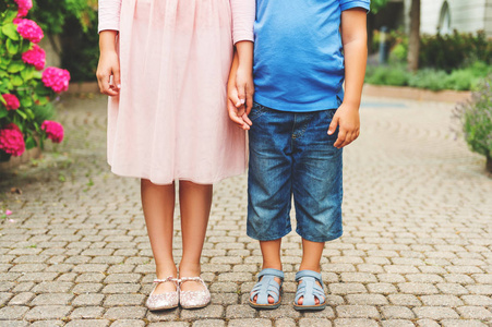 两个成对的小孩脚穿时装鞋，小男孩穿着牛仔短裤和蓝色皮革凉鞋，女学生在粉红色的连衣裙和芭蕾舞鞋