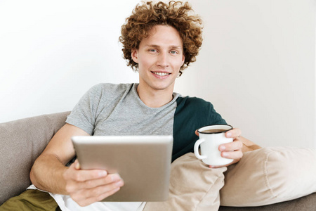英俊的男人微笑着坐在沙发上使用平板电脑