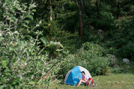 在山中露营。一个女人坐在附近的绿树和山背景下的帐篷