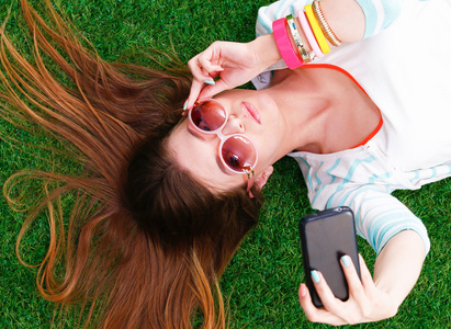 美丽的年轻女子躺在绿草通过她的手机拍照
