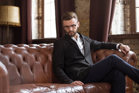 英俊的时尚自信胡子商人在一个聪明的西装和眼镜坐在沙发上办公室