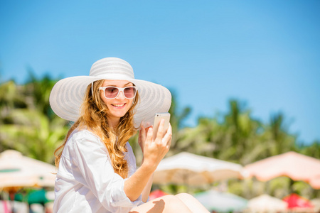 美丽的年轻女子在沙滩上坐在阳光明媚的日子，在她手中的手机
