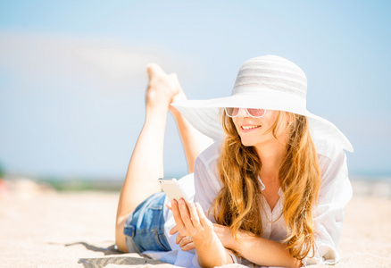 美丽年轻女子在沙滩上躺在阳光明媚的日子，在她手中的手机