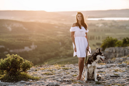 漂亮的女孩在日落时玩一只狗 黑色和白色哈士奇蓝眼睛 在山上