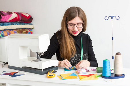针线活和车间的一位年轻女子，一个裁缝年轻在眼镜裁缝在工作线程 面料 针与缝纫机桌上的彩色布片绗缝