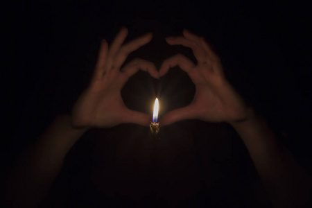 在完全黑暗的背景下创建的背后是燃烧的蜡烛的爱心形状的女性手