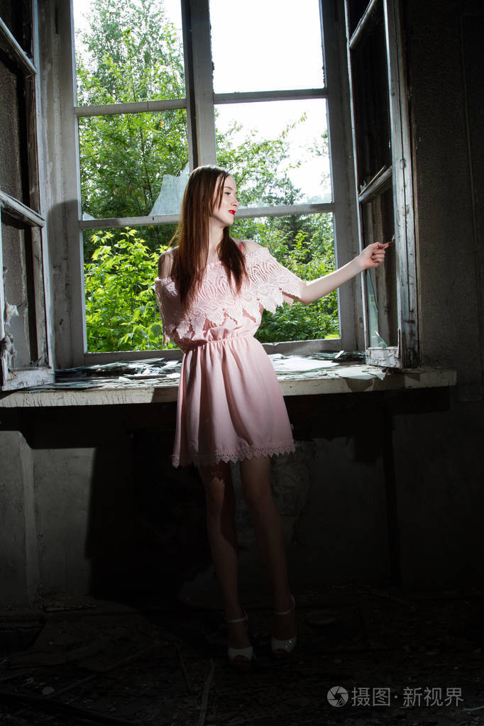 穿着粉红色的短连衣裙的女孩正站在一个废弃的房子的窗前