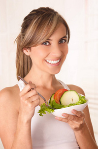 美丽的年轻女子，穿着白色 t 恤和她手里拿着一份健康的沙拉和使用叉子