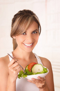 美丽的年轻女子，穿着白色 t 恤和她手里拿着一份健康的沙拉和使用叉子