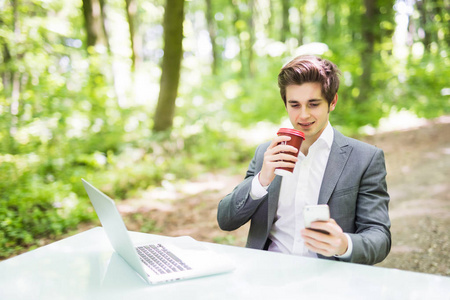 商人与便携式计算机和杯咖啡坐在办公室桌前使用手机在绿色的森林公园。经营理念