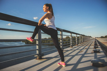 拉伸后锻炼。横向拍摄的美丽的年轻女子在体育穿看着别处做拉伸在桥上了蓝蓝的天空背景上的时候