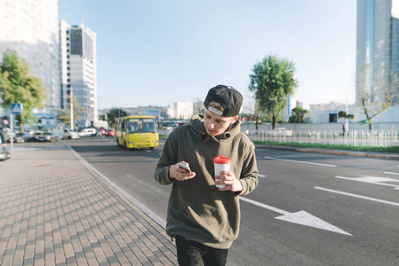 一个时尚的年轻人，与一杯咖啡在他的手沿着人行道和使用手机。一个学生写到消息上的道路和汽车背景