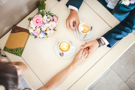 新婚夫妇在咖啡馆，顶视图。男人握着女人的手，喝咖啡。新娘和新郎喝咖啡休息时间约会礼物，桌上的花束