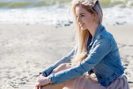 在海边的年轻开朗的女孩。年轻的金发女人坐在沙滩上。时尚米色裙子
