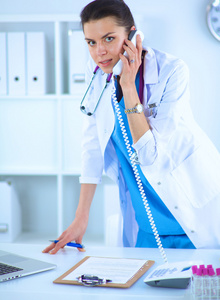 年轻的女医生穿白色上衣，在计算机上使用电话的肖像