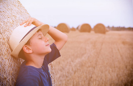 与草帽子可爱的乡村男孩在附近草垛在日落时在夏天的帽子.