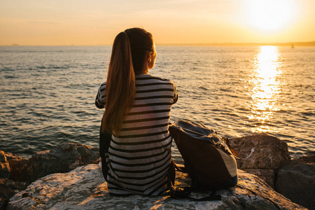 年轻游客的女孩背着日落时分坐在海边岩石上，眺望远方。休息 放松 旅行 度假