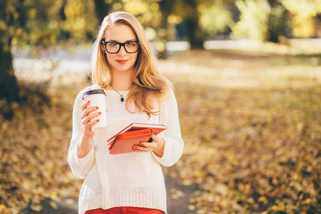 年轻金发女学生戴眼镜, 长头发捧着咖啡, 在公园户外看书秋季时间教育, 生活方式和人的概念