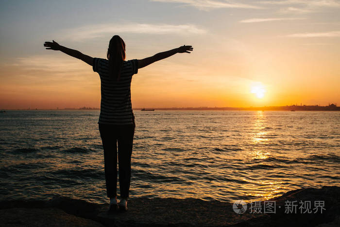 从后面的年轻女孩举起双手靠在海边日落背景.自由, 休息, 旅游, 度假.