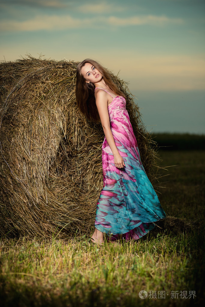 年轻的女子站在晚上场在干草堆里。时尚麦粒肿