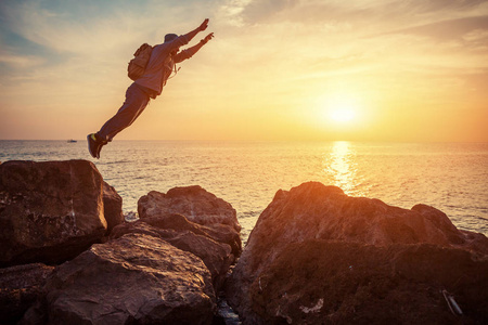 年轻勇敢的男子跳过海边岩石在日落，背包里