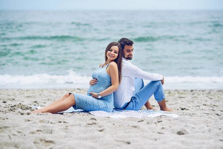 美丽的孕妇和她互相依偎，坐在海边的正派人。可爱的年轻夫妇