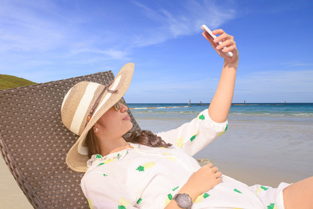 美丽的亚洲女孩自拍照与智能手机在夏季海滩附近的长椅上