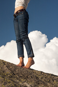 女人的腿 脚和手与牛仔裤，在岩石上，与自然背景和蓬松，白色的云