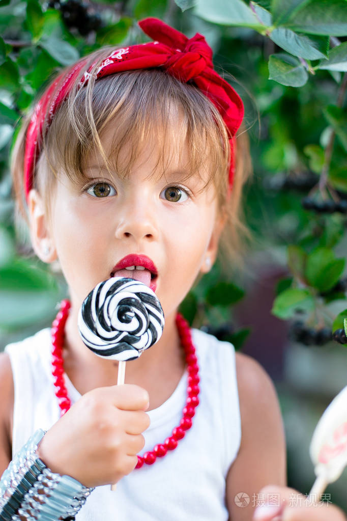 很酷的小女孩舔着棒棒糖室外孩子在白色t恤牛仔短裤红色项链和头上的