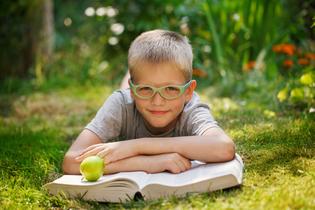 可爱的小宝贝男孩戴着眼镜躺在绿色的草地，在夏季的一天读这本书