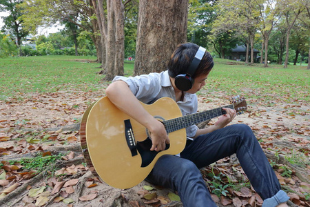 快乐的年轻人玩吉他，斜倚在室外公园一棵树的画像