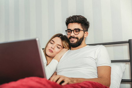 年轻幸福夫妇躺在床上, 在家里使用笔记本电脑