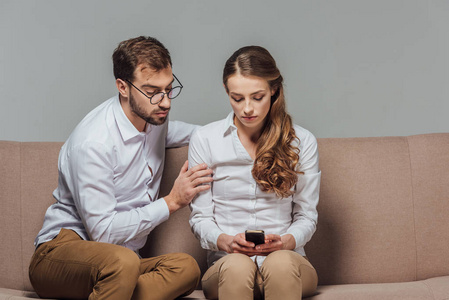 严重的年轻女子使用智能手机, 而英俊的男子在眼镜坐在沙发上的灰色隔离