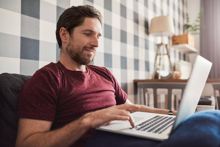 微笑的年轻人在他的客厅沙发上独自坐在家里用手提电脑浏览互联网