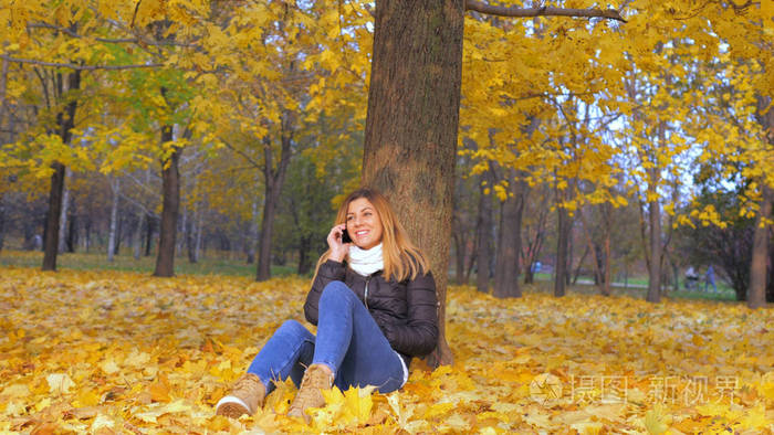 女人坐在他的背部到树在黄色的秋天树叶, 说话智能手机