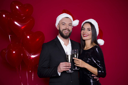 有一个美好的新年和圣诞节。欢快的美丽微笑情侣在圣诞老人的帽子与香槟在红色的背景与球的眼镜。情侣度假