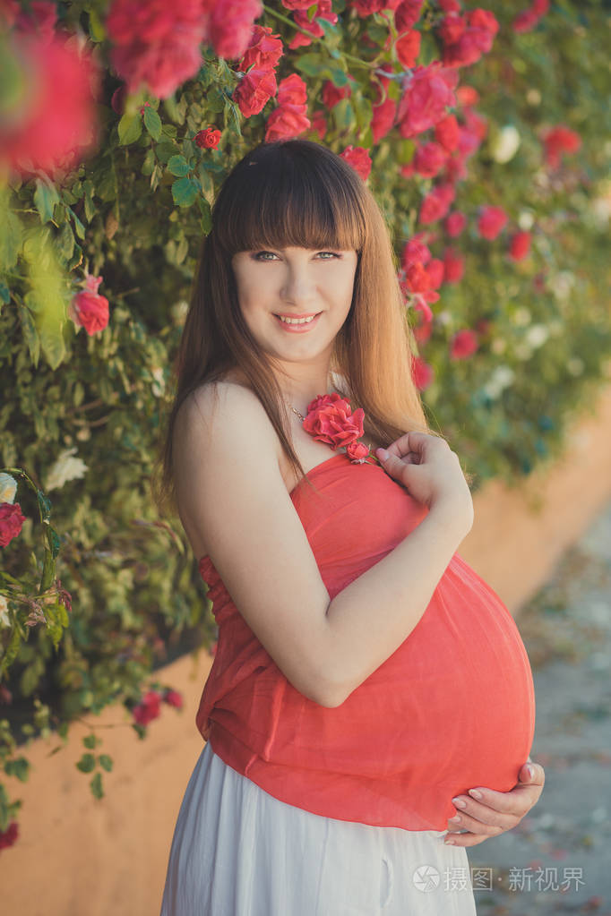可爱的怀孕的年轻妈妈穿着轻快的红色白衣装扮着夏日里的红桃玫瑰在
