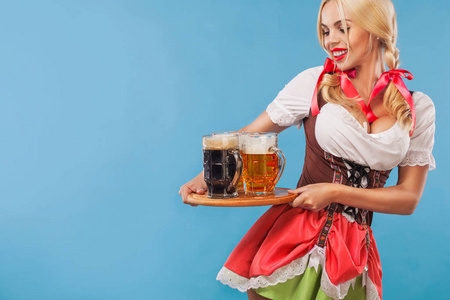 年轻慕尼黑啤酒节服务员，穿连衣裙的女孩传统巴伐利亚，服务在蓝色背景上的大啤酒杯