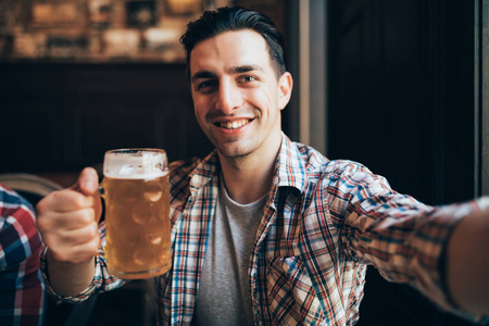 一个兴奋的年轻人尖叫着高兴地拿着一杯啤酒在当地的酒吧放松自拍