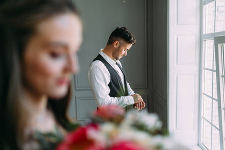 英俊的新郎准备婚礼背景模糊新娘的花束上。在靠近全景窗户的节日衣服的男人。图稿