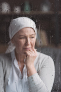 病成熟妇女在头巾哭泣在窗口后面与雨滴