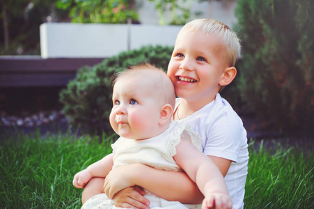 快乐的小兄弟玩拥抱他的妹妹婴儿坐在草地上的绿色花园, 爱和养育的概念