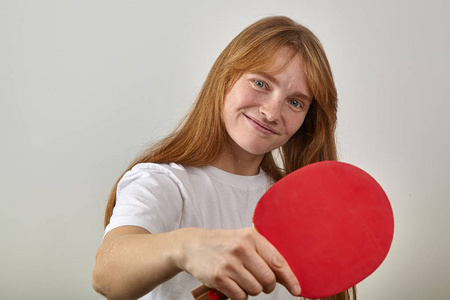 年轻妇女与红头发和雀斑身着白色 tshirt 手持乒乓球拍和看着相机