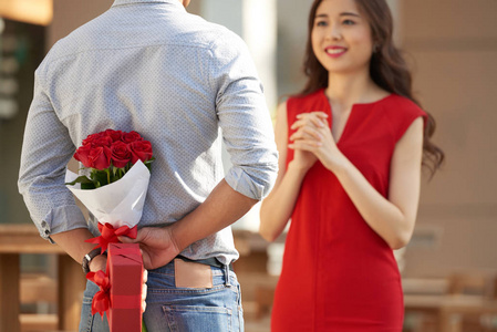 美丽的亚洲女人穿着红裙子看着她的男友, 男人藏在他背后的红玫瑰花束和礼品盒