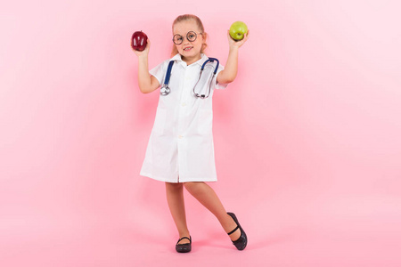 可爱的小女孩在医疗制服与听诊器和二个苹果在粉红色背景