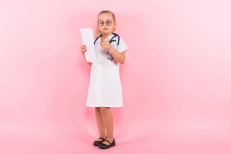 可爱的小女孩在医疗制服与听诊器, 白色床单和钢笔在粉红色背景
