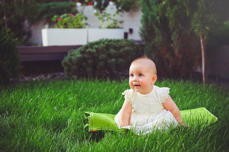 一个半岁的孩子坐在院子里的草地上, 穿着一件白色的连衣裙, 欣喜地, 6 月。儿童儿童用品的概念教育