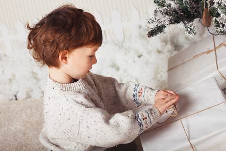 可爱的小男孩正坐在圣诞树旁的礼物上。这个孩子对新年礼物很满意。白种金发小孩